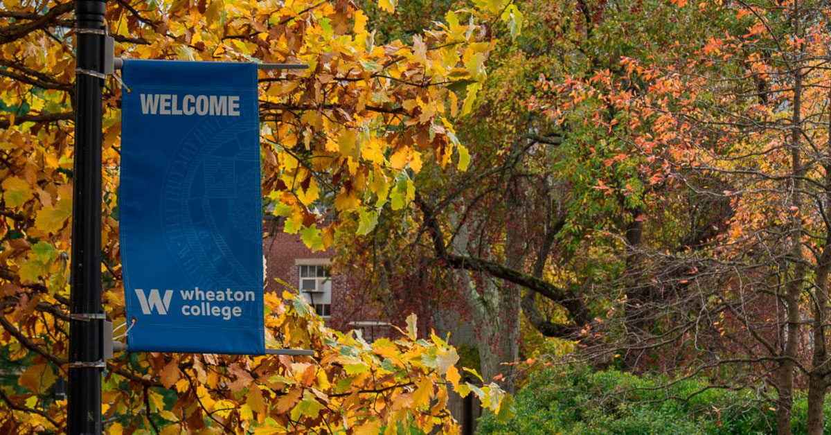 A blue banner reading Wheaton College is attached to a black post with colorful foliage in the background