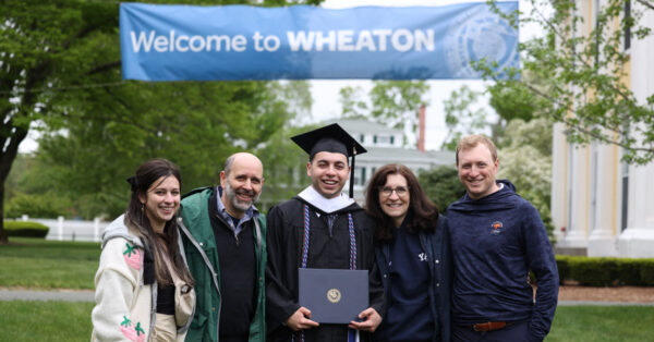 A graduate poses for a photo with family and friends under the Welcome to Wheaton banner.