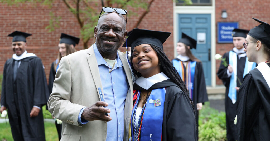 A graduate poses for a photo with their parent. Both are smiling happily at the camera.