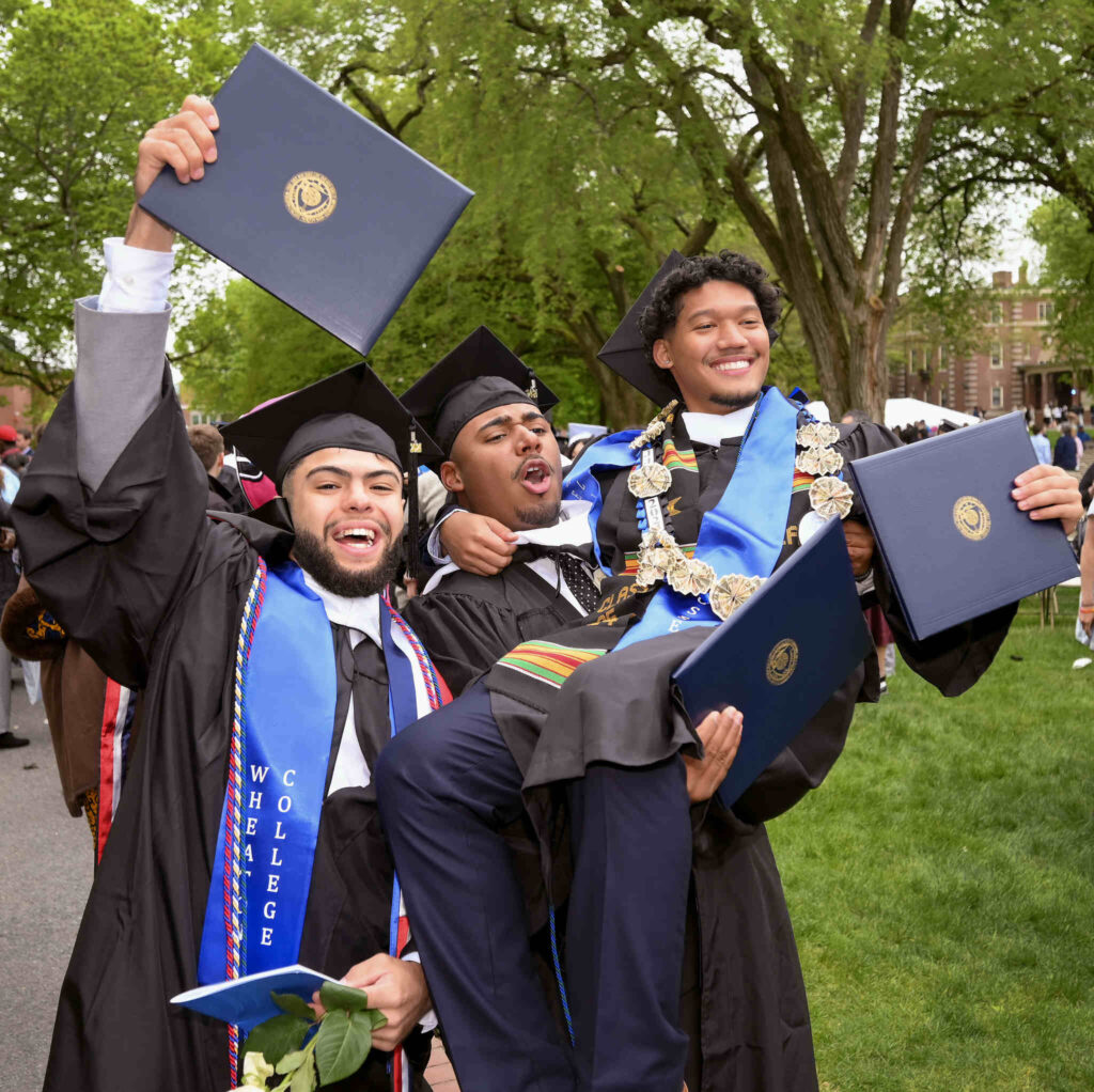 A group of three graduates celebrating joyously, one of them lifting the other up in their arms.