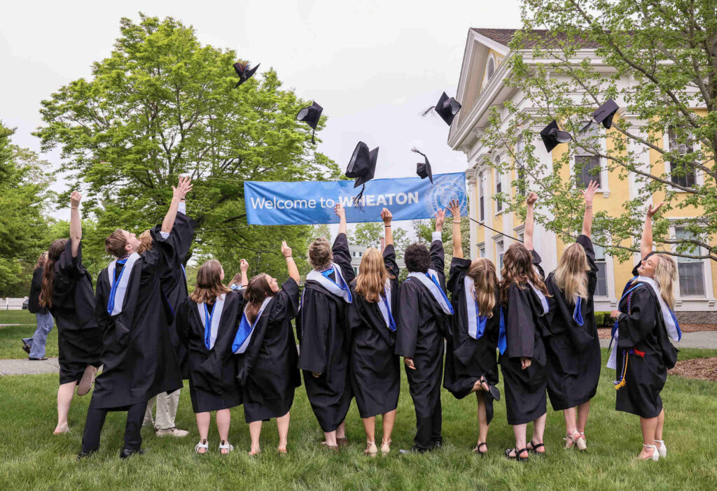 A group of graduates throw their caps up into the air under a Welcome to Wheaton banner.