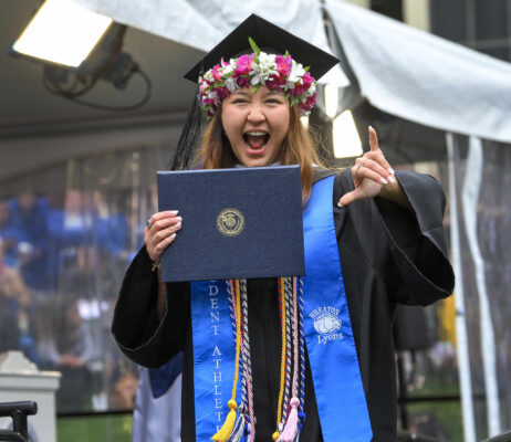 A happy graduate holds up their diploma and gives the camera a thumbs up.