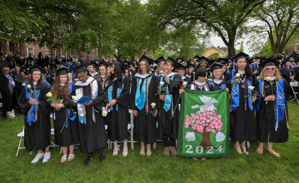 Class of 2024 graduates stand at the beginning of their Commencement ceremony. Their class flag, a brightly embroidered tree, is front and center.