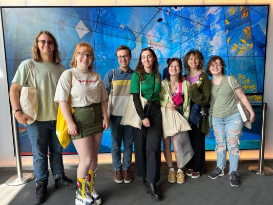 Six individuals smile in front of a large painting at a Frameless Art Exhibit.