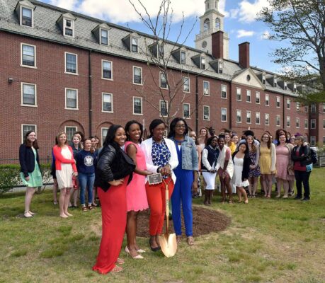 A group of young people stand in a courtyard next to a newly planted tree, with one person holding a golden shovel used for the ceremony.