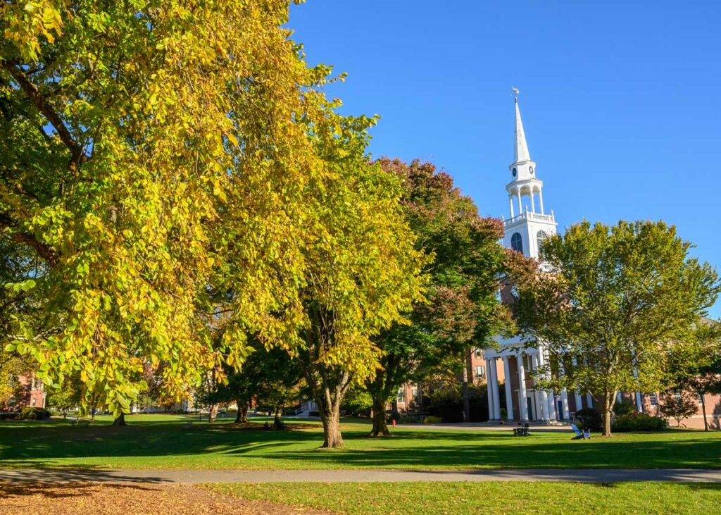 A grove of mature trees with leaves turning yellow, gold, orange and red stand with a chapel steeple in the background and a blue sky above.
