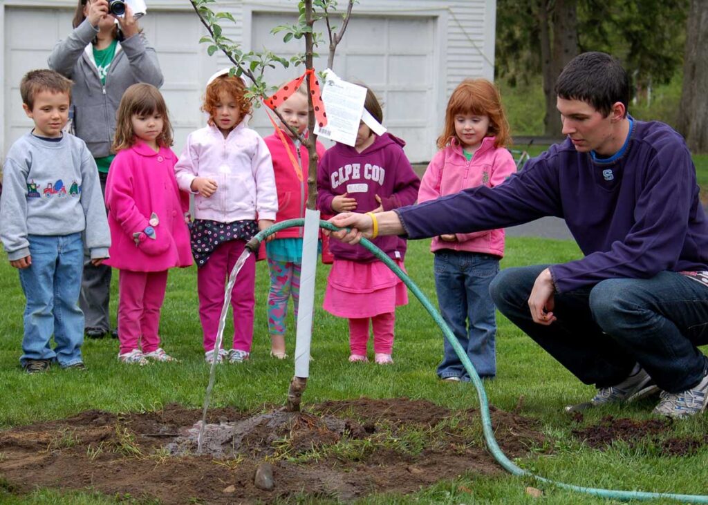 A group of young children watch as a young man waters an apple sapling.